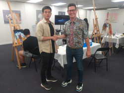 Edward shaking hands with Evan, standing in front of student artists, who are painting on easels in the background