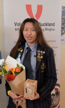 Aira standing in front of a banner for Volunteering Auckland. Holdiong a bunch of flowers and a Keep Cup that were gifts for presenting at the Love Awards 2022