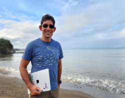 Urs is wearing blue t shirt standing at a beach holding his laptop